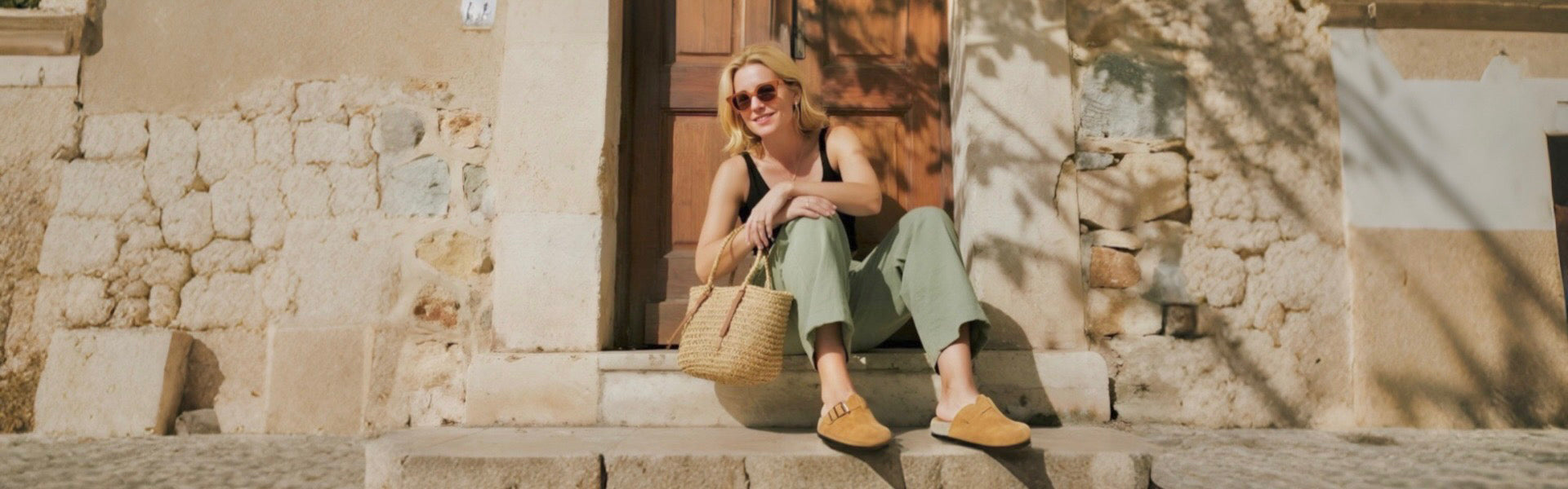 A woman sitting on stone steps wearing light green pants and mustard yellow Birkenstock sandals, holding a woven straw bag on a sunny day