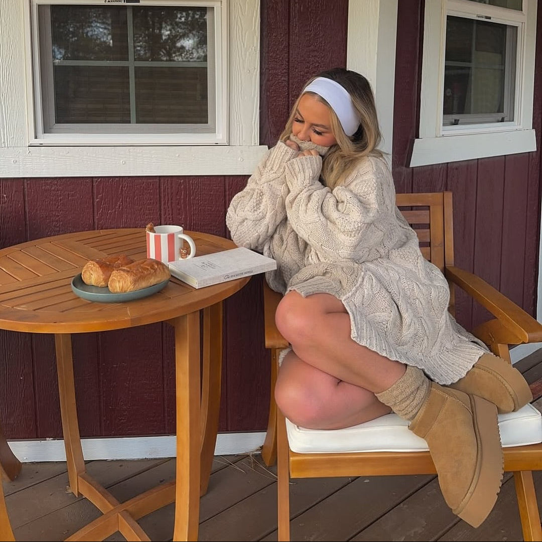 A young woman sitting comfortably on a wooden porch chair, wearing a chunky cream cable-knit sweater dress, a white headband, and tan platform mini boots