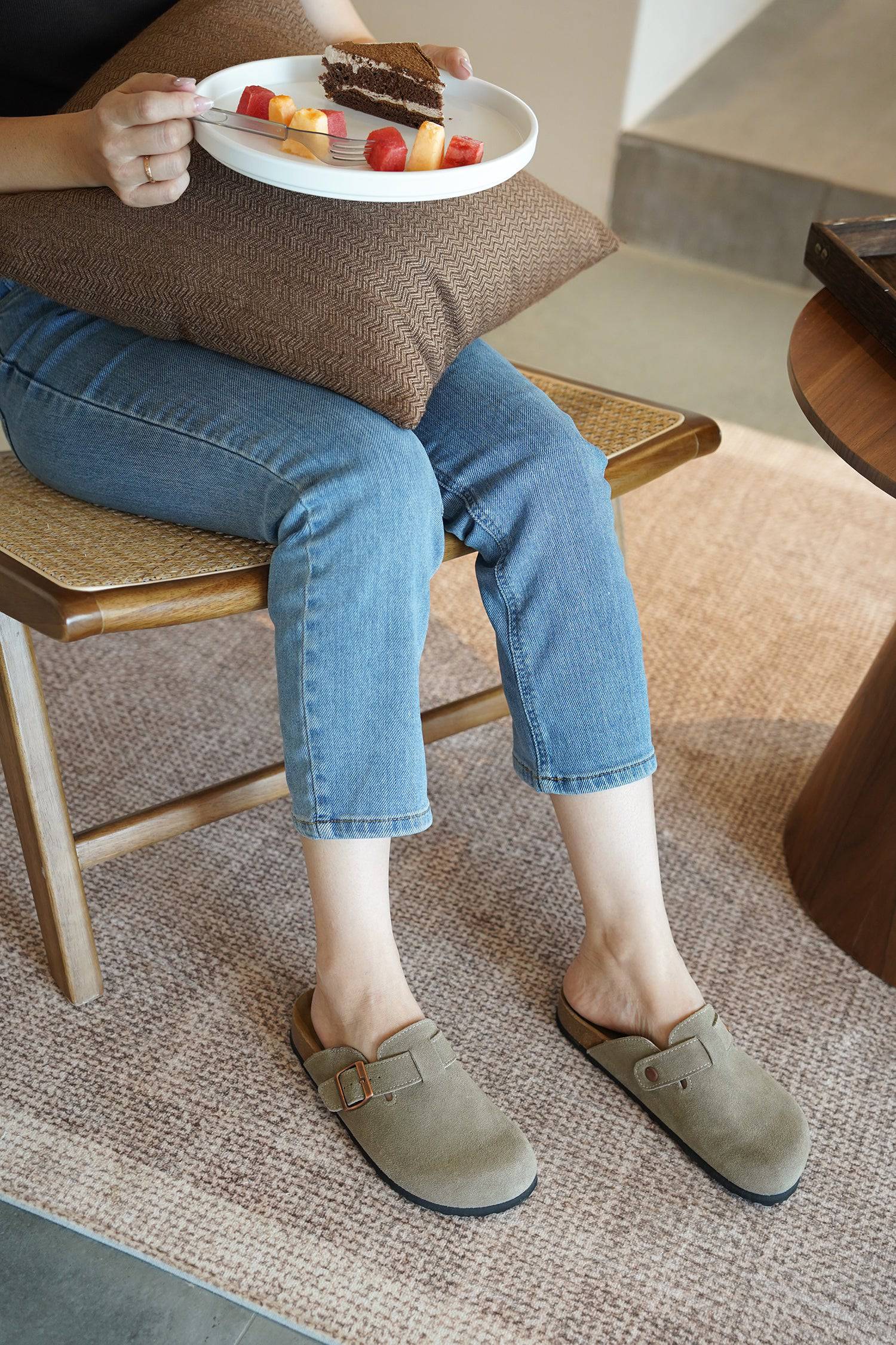 Woman wearing Project Cloud Benton suede clogs, resting on a chair while holding a plate of dessert.